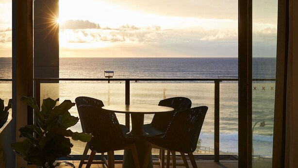 A balcony with a view of the ocean at sunset, featuring wicker chairs and a small table.