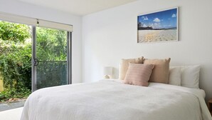 A bedroom with a large bed, a view of greenery through a sliding glass door, and a beach scene on the wall.