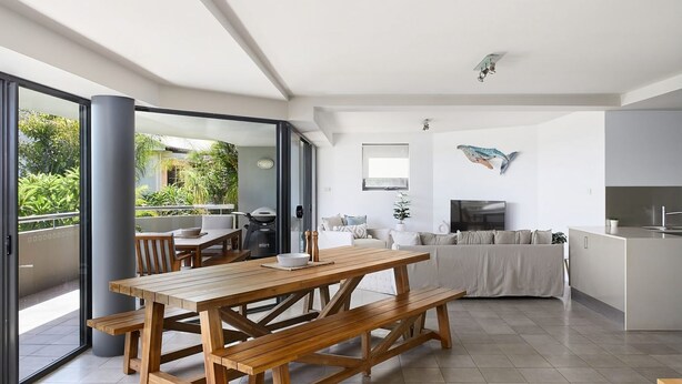 A modern living room with a wooden dining table, a sofa, and a TV. There is a balcony with a grill and plants visible through the glass doors.