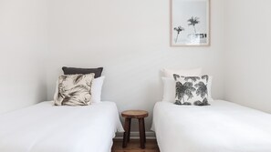 Two single beds with white bedding and decorative pillows, a small wooden stool, and a framed picture on the wall.