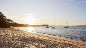 Ein Strand mit in ruhigem Wasser vor Anker liegenden Booten, einem klaren Himmel und einer Sonne, die am oder über dem Horizont untergeht bzw. aufgeht.