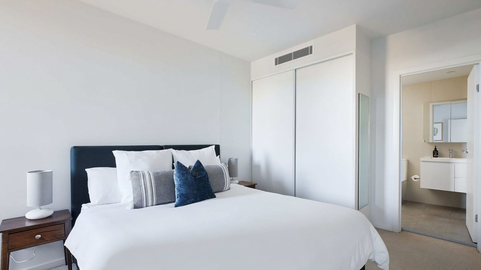 A modern hotel room with a large bed, a nightstand, a bedside lamp, a wardrobe, and a bathroom visible through an open door.