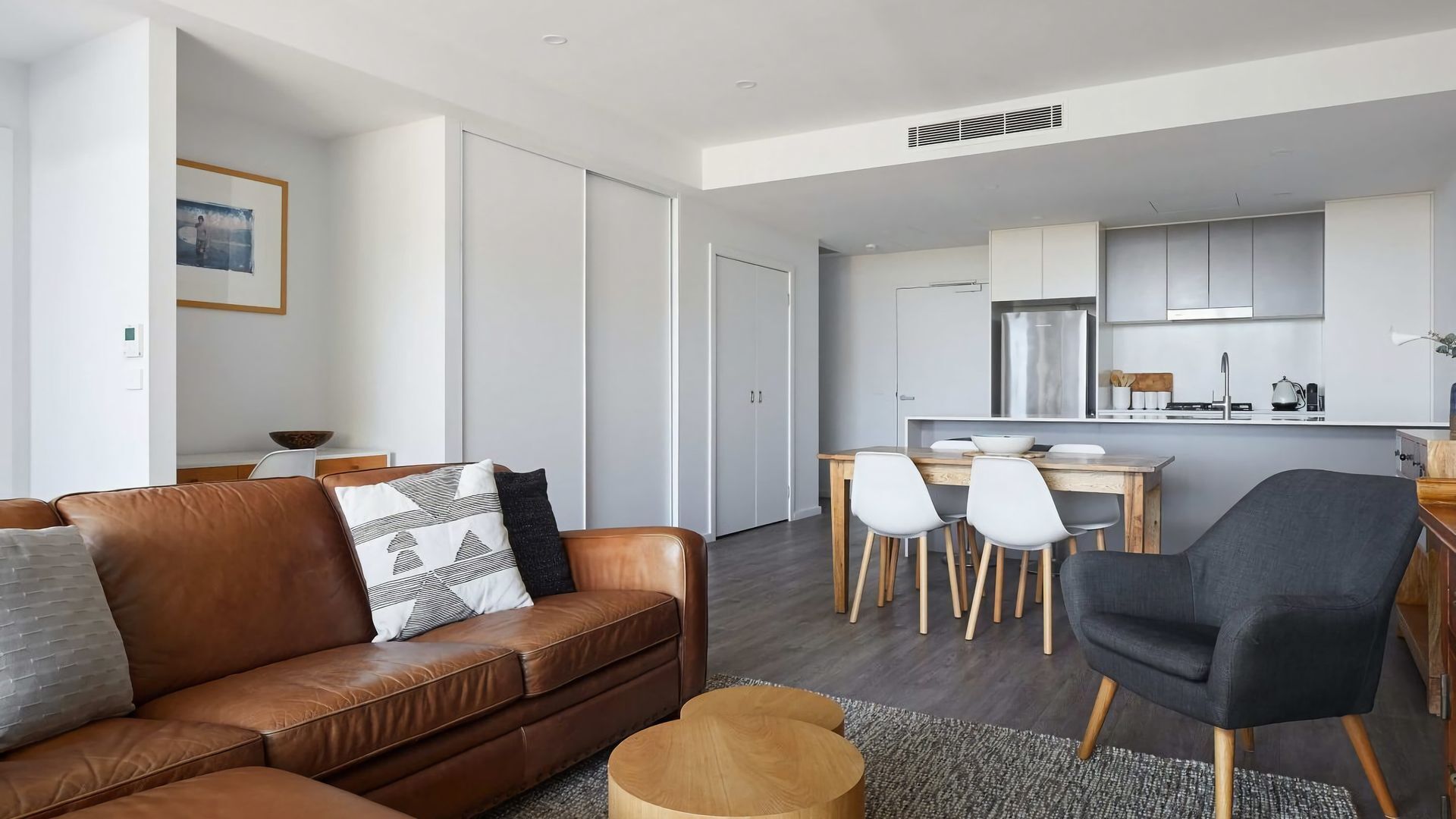 A modern living room with a brown leather sofa, a black armchair, and a dining area with a wooden table and white chairs.
