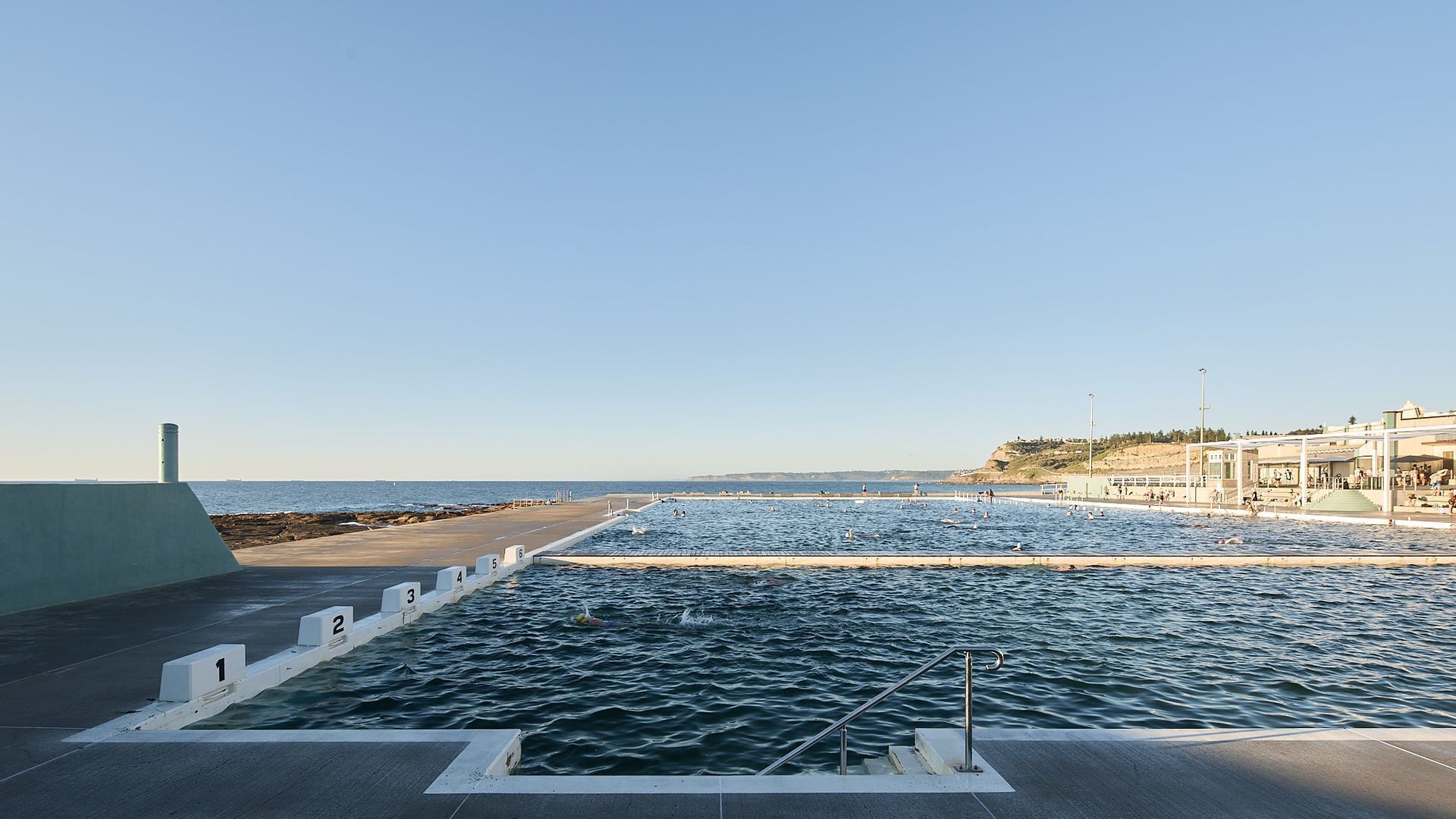 A swimming pool with numbered lanes, a viewing deck, and a beach in the background.