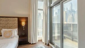 Una habitación de hotel con un ventanal grande, una cama con ropa de cama blanca y una mesita de noche con lámpara.