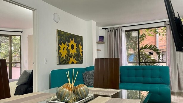 A modern living room with a teal sofa, a glass coffee table, and a painting of sunflowers. - Exclusivo Apartamento Privado Zona Rosa Sabaneta (Sabaneta)