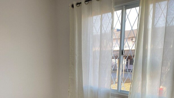 A dining area with a wooden table and four chairs, a window with a view of buildings, and a curtain. - Residencial Karine - Studio 05 (Joinville)