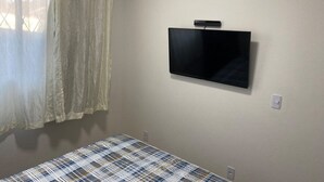 A room with a bed, a television mounted on the wall, an air conditioner, and a window with curtains. - Residencial Karine - Studio 05 (Joinville)