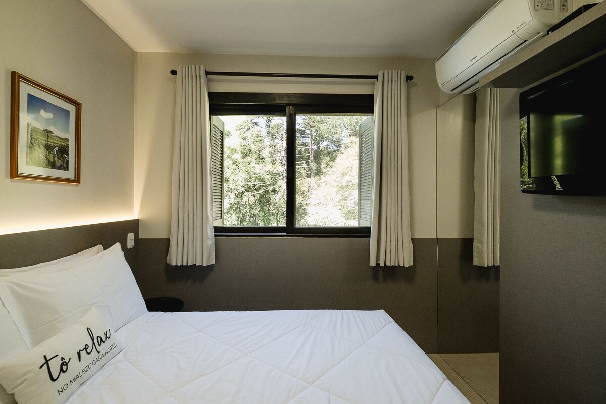 A hotel room with a bed, a window with curtains, an air conditioning unit, and a television on the wall.
