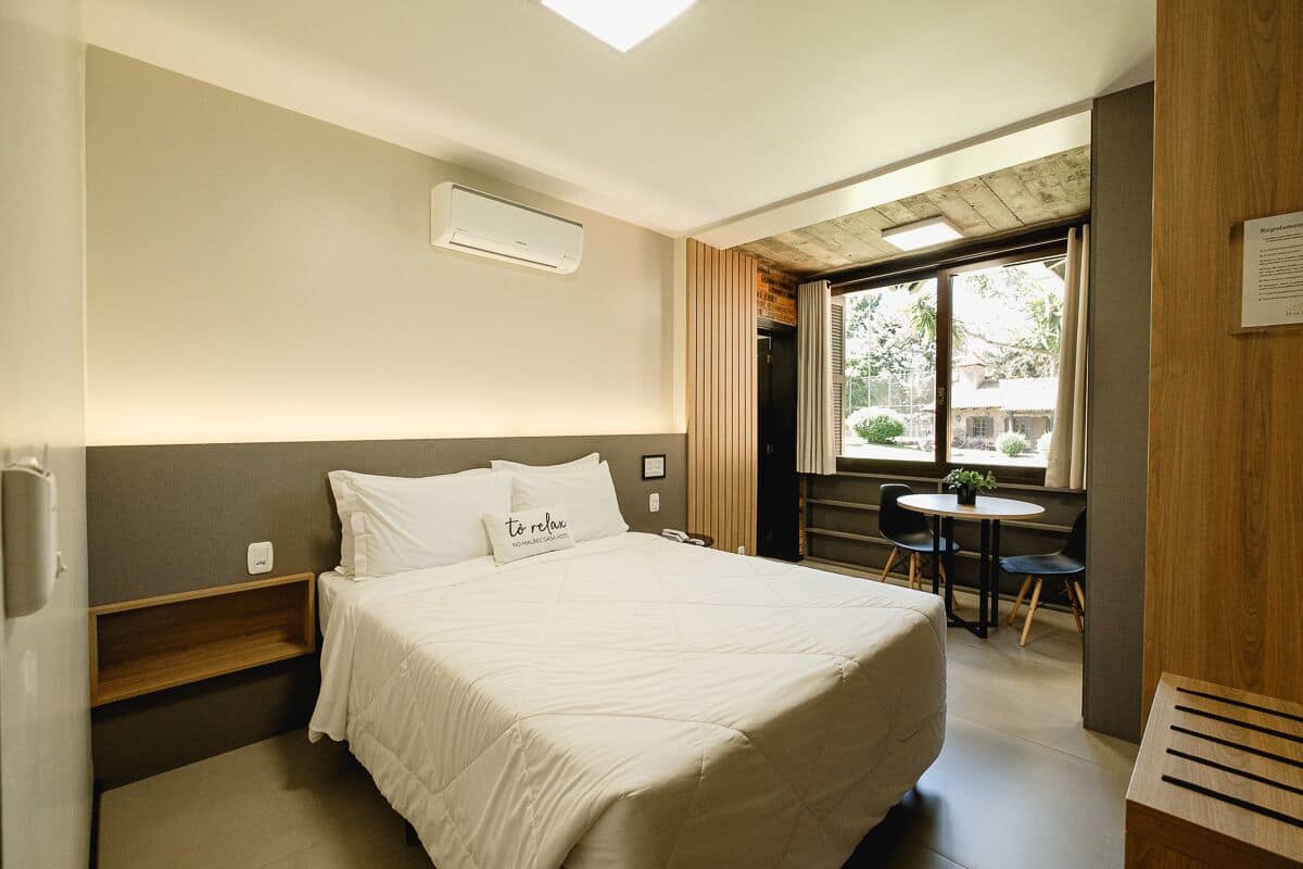 Hotelkamer met een bed, nachtkastje en een klein bureau met stoel. Er is een raam met gordijnen, een plant op het bureau en een wandairconditioner.