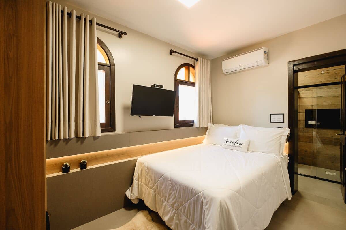 Een slaapkamer met een bed, een televisie, een raam met gordijnen, een wandairconditioner en een glazen deur naar een douchegedeelte.