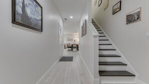 A hallway with a staircase, framed artwork on the walls, and a dining area in the background.