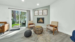 A modern living room with a fireplace, two armchairs, a round coffee table, and a large window overlooking a garden. - Huge CDA Getaway With Foosball And BBQ (Coeur d'Alene)