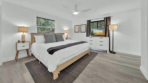 A modern bedroom with a large bed, bedside lamps, a ceiling fan, and a view of a sunset through a window. - Huge CDA Getaway With Foosball And BBQ (Coeur d'Alene)