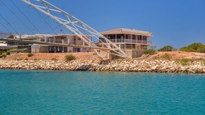 A coastal building with a balcony overlooking turquoise waters, a rocky shoreline, and a bridge structure in the background. - 120 Madaffari Drive - Featuring two master suites and a full outside bathroom (Exmouth)