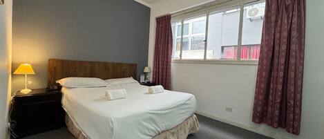 A neatly made bed with a wooden headboard, a bedside table with a lamp, and a window with patterned curtains.