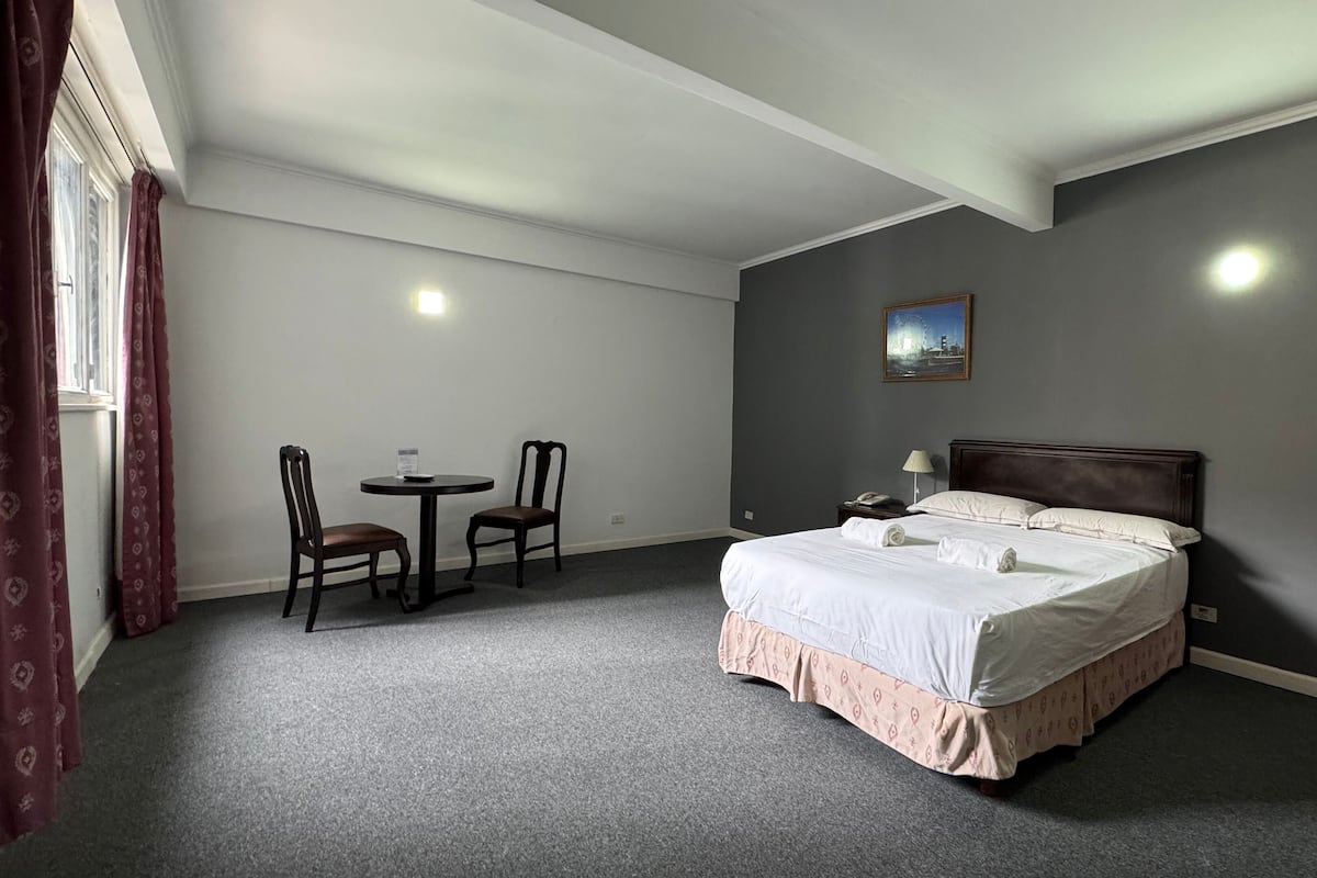 A hotel room with a bed, a small table with chairs, a nightstand, and a painting on the wall.