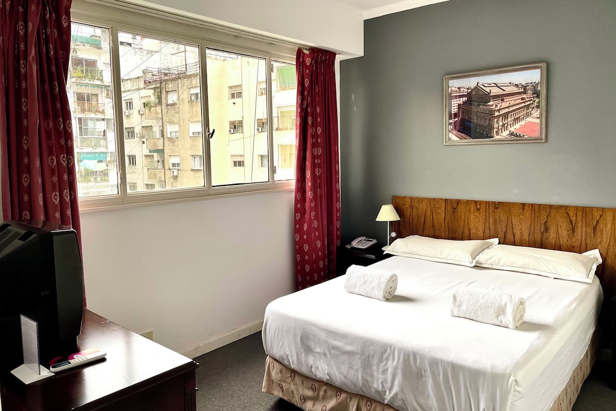 A hotel room with a bed, a desk with a TV, a phone, and a window with curtains.