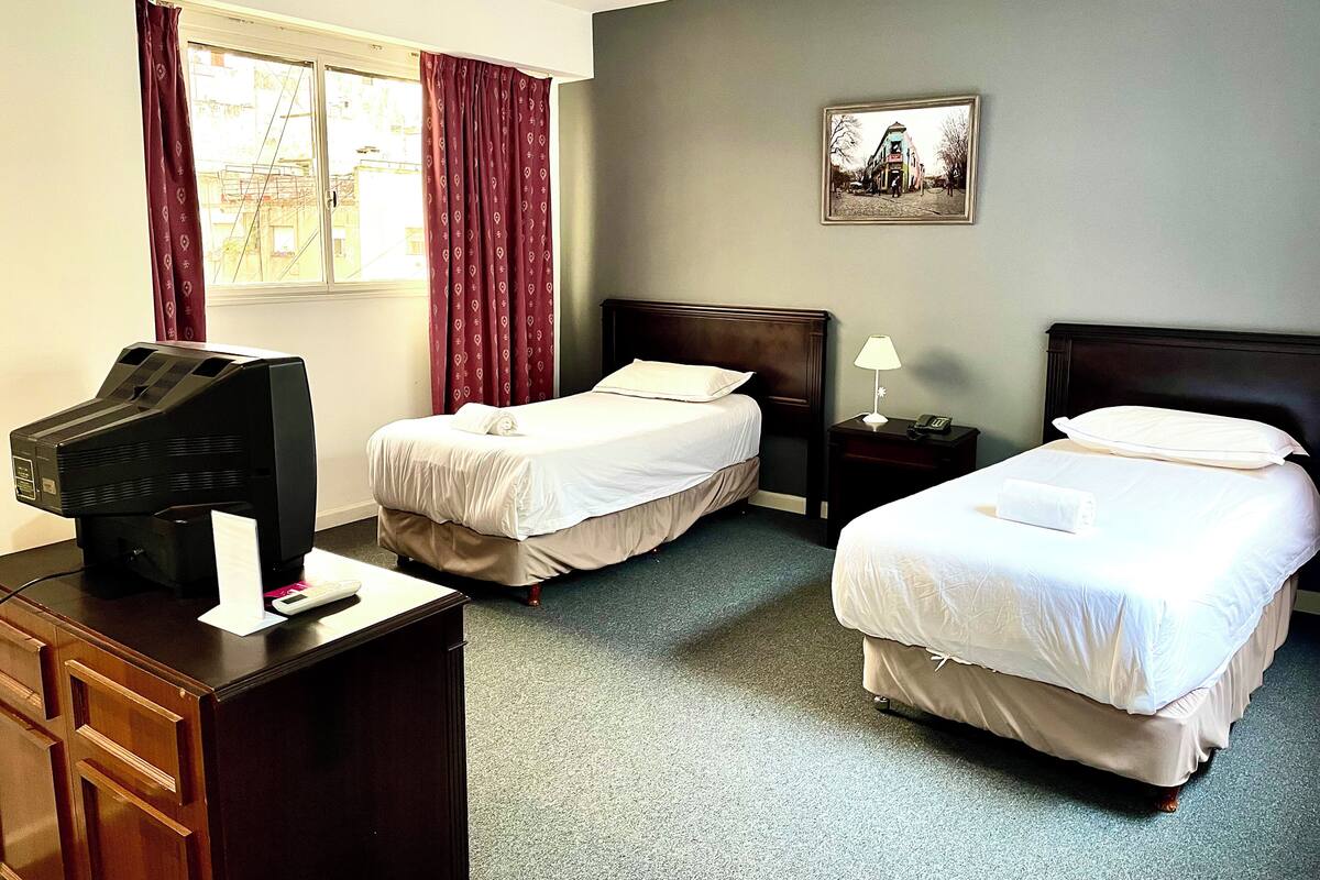 A hotel room with two single beds, a desk with a television, a nightstand with a lamp, and a framed picture on the wall.