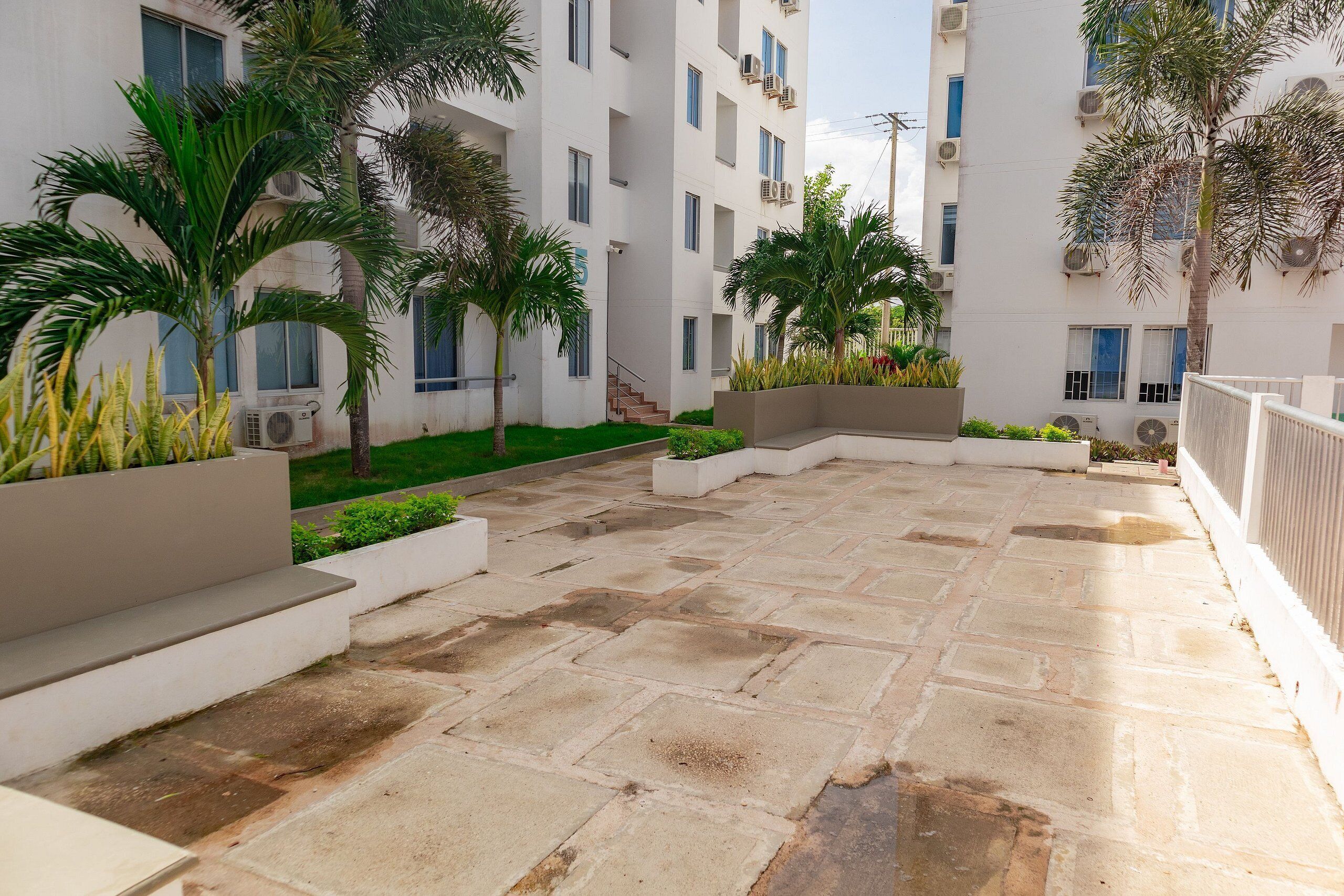 A courtyard with stone flooring, palm trees, and a white building with multiple windows and air conditioning units.