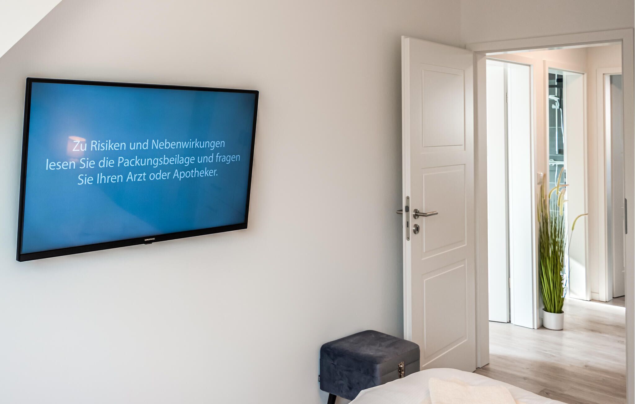 A modern bedroom with a large flat-screen TV mounted on the wall displaying a message in German. There is a white door leading to a balcony with a potted plant.
