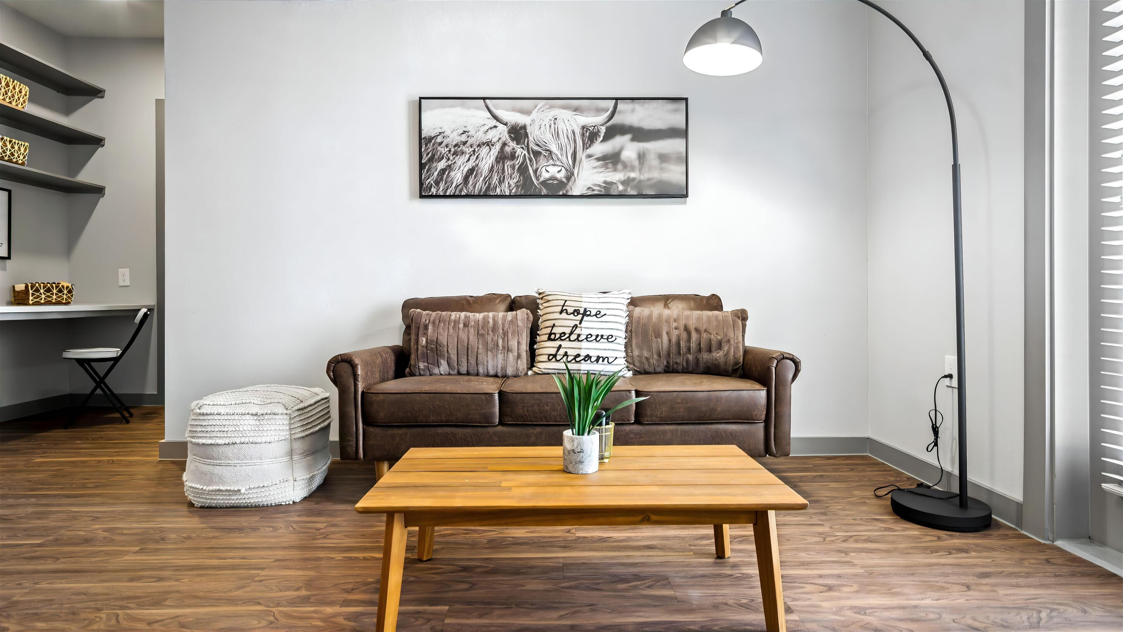 A modern living room with a brown leather sofa, a wooden coffee table, and a black floor lamp. A framed black and white photograph hangs on the wall above the sofa.
