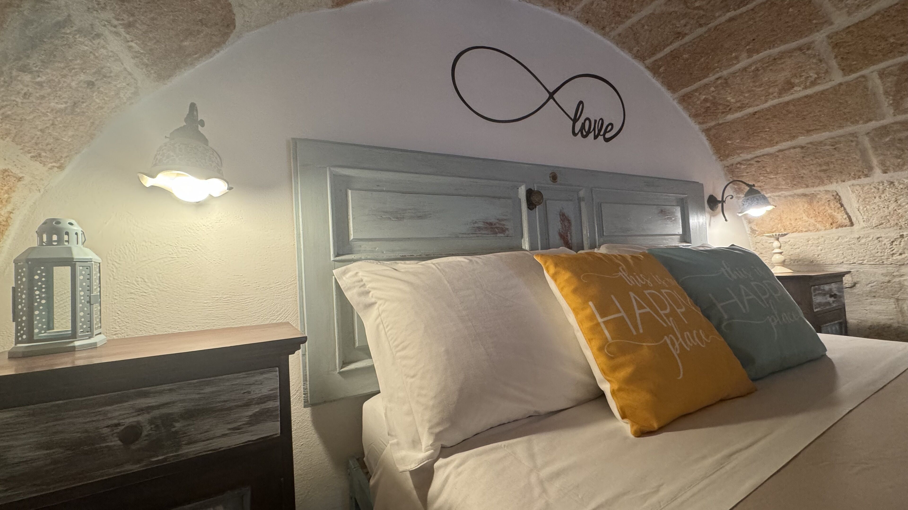 Quarto com cama, mesa de cabeceira, um candeeiro e uma parede com a inscrição 'love'.