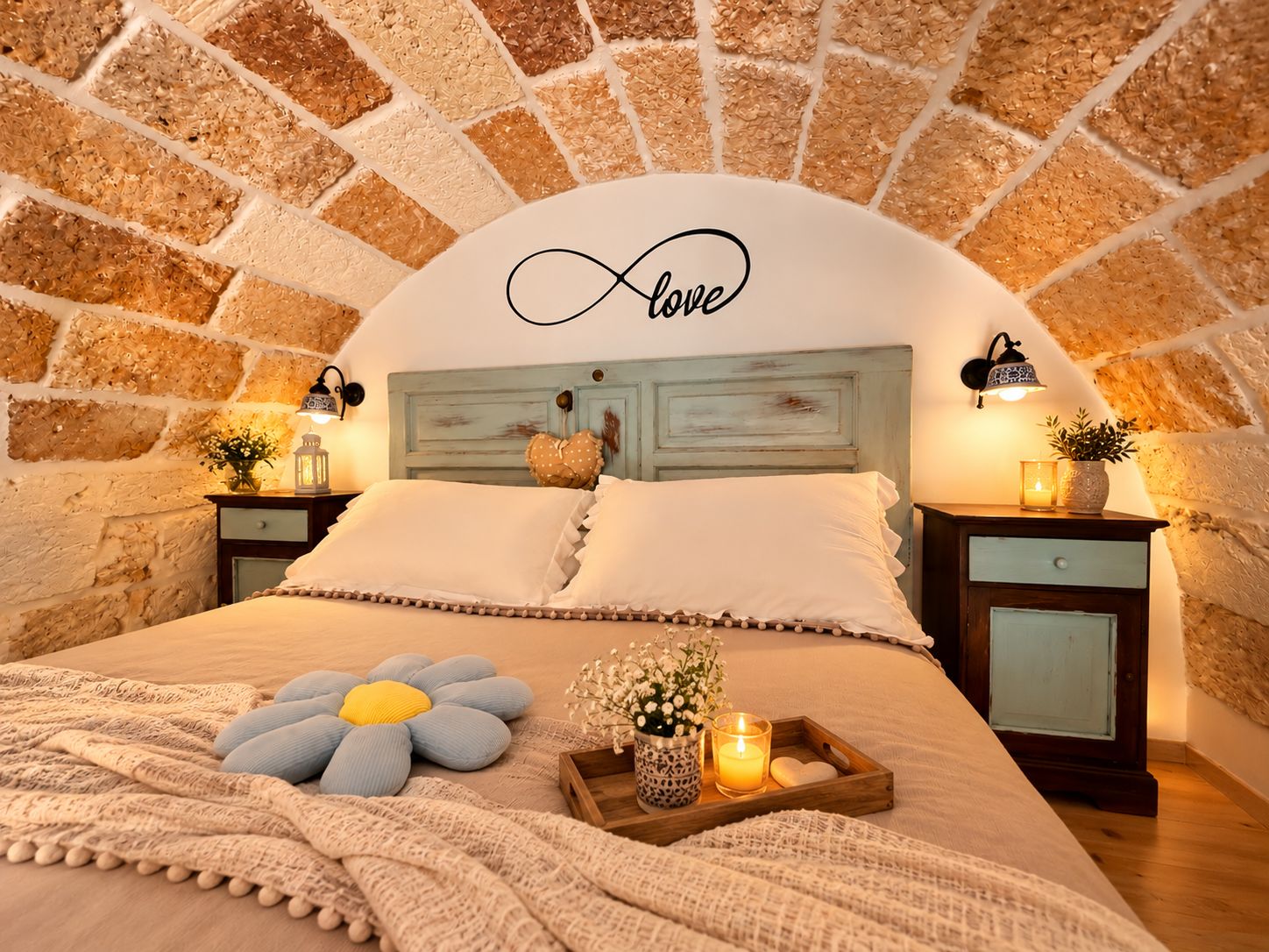 Een slaapkamer met een stenen muur, een bed met wit beddengoed, een decoratief oneindigheidssymbool en een nachtkastje met een lamp.