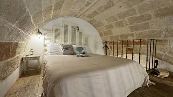 Un dormitorio con techo de piedra, una cama con ropa blanca, una mesita de noche con un farol y una ventana con contraventanas.