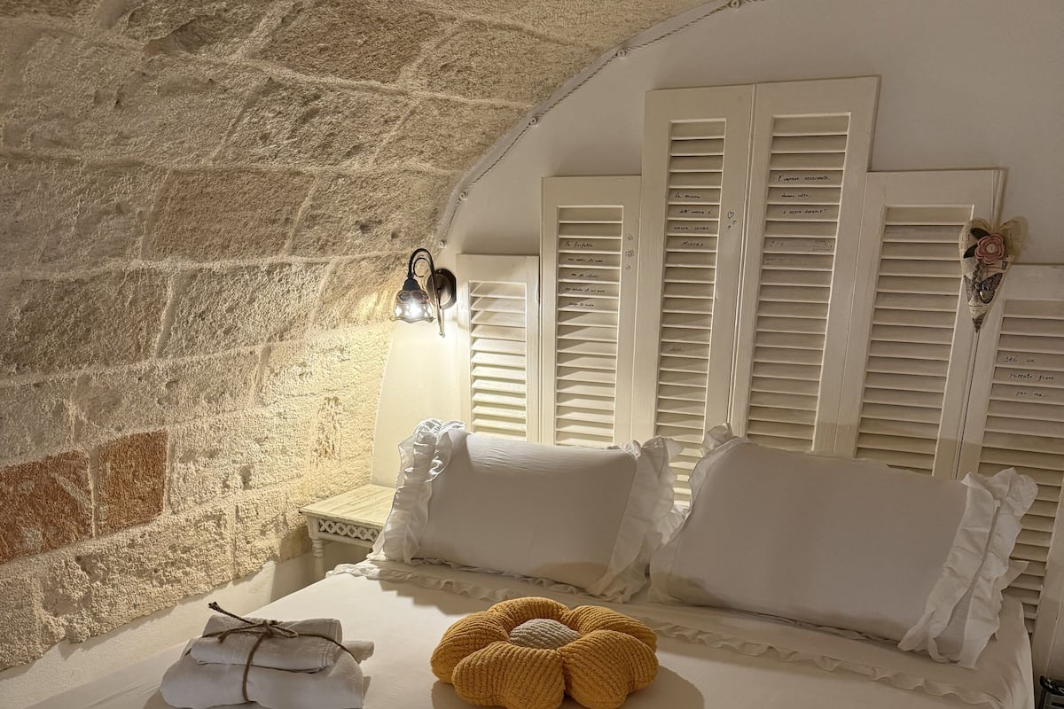 Un dormitorio acogedor con pared de piedra, cama con ropa blanca, una flor amarilla tejida y una mesita de noche con lámpara.