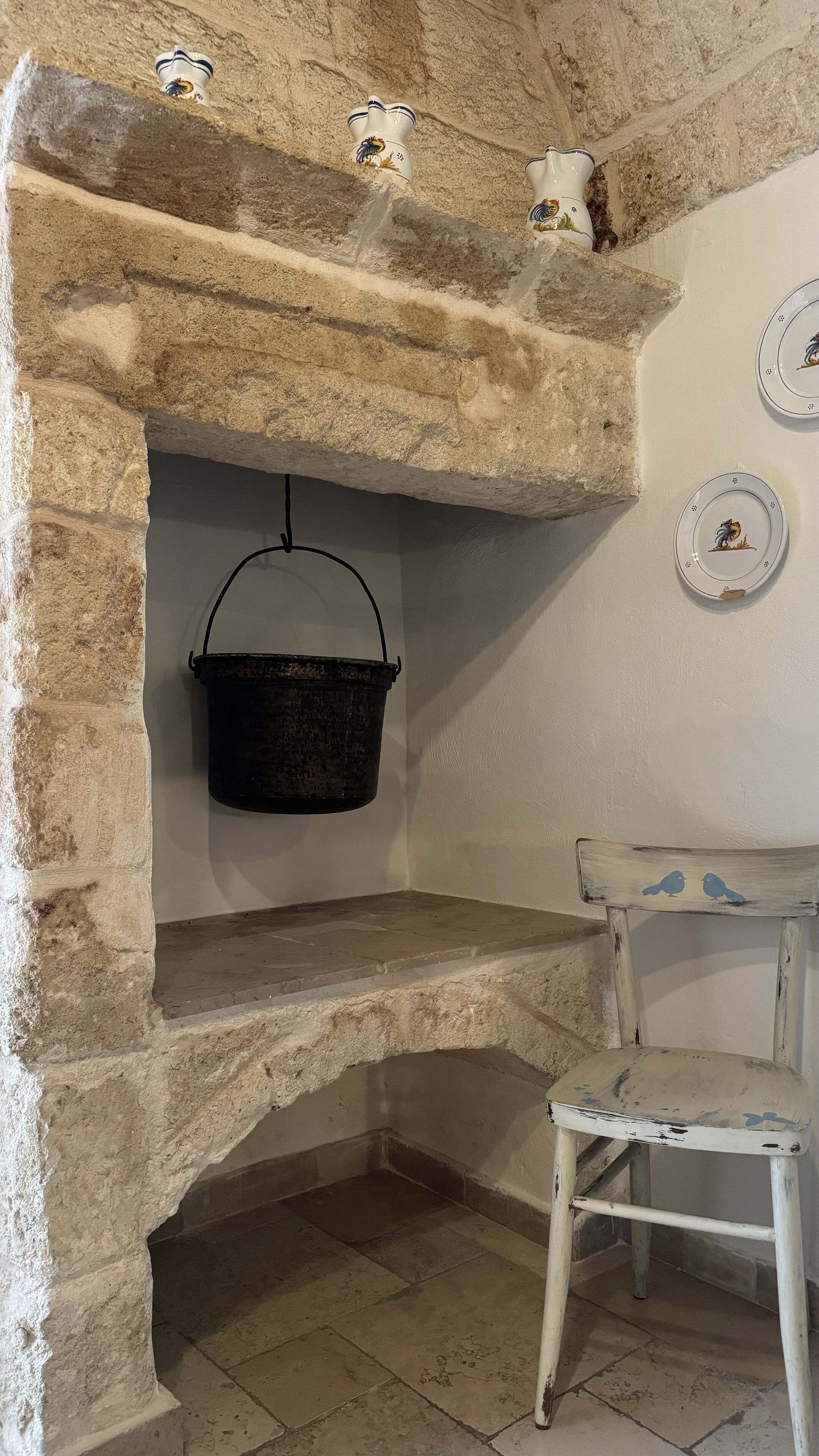 Una chimenea de piedra rústica con un cubo metálico colgante, una silla pintada de blanco y platos decorativos en la pared.