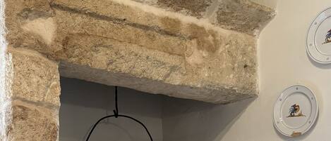 Una chimenea de piedra rústica con un cubo metálico colgante, una silla pintada de blanco y platos decorativos en la pared.