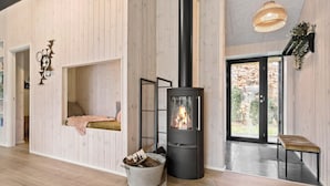 A modern interior with a wood-burning stove, a seating area with cushions, and a large window offering a view of the outdoors. - 4 bedroom gorgeous home in Glesborg (Glesborg)