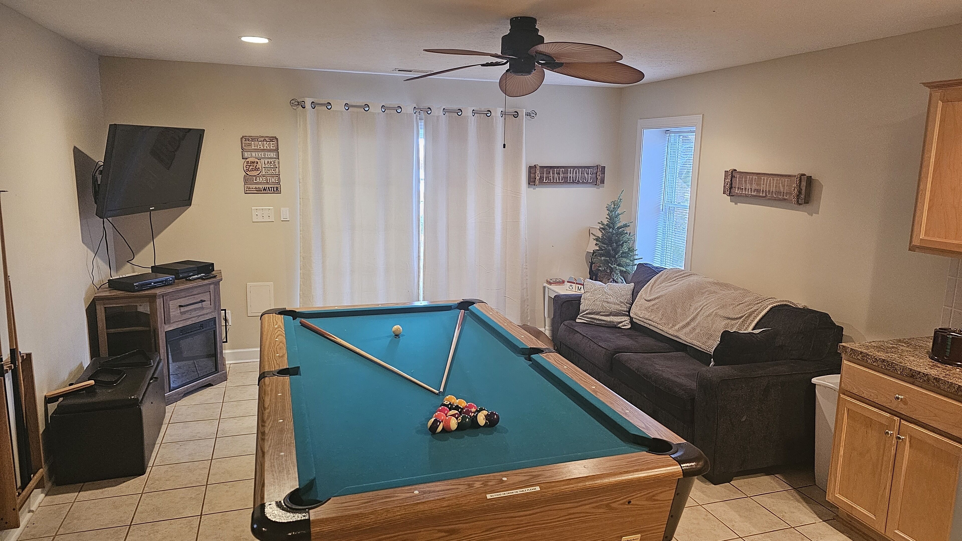 A pool table in a living room with a ceiling fan, a television on a stand, a couch, and a window with curtains.