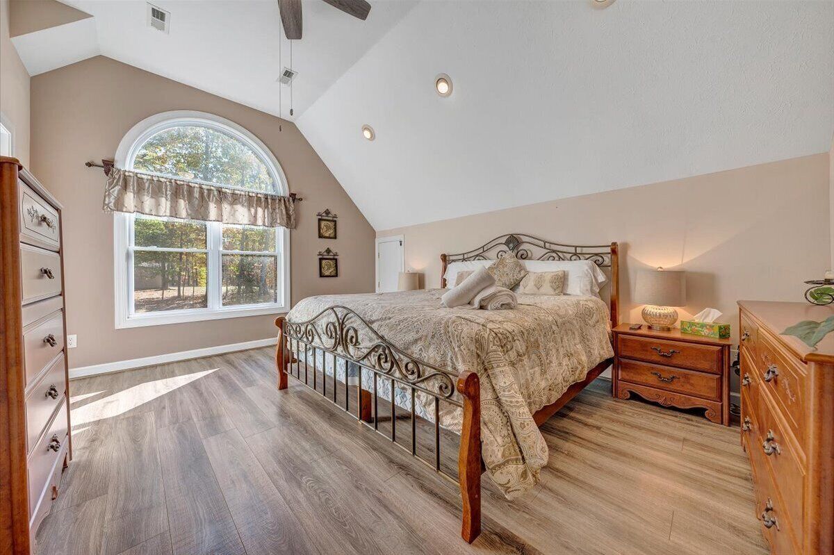 A bedroom with a large bed, a dresser, a nightstand, and a window with a view of trees.