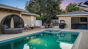 Un patio trasero con piscina, una zona de estar con una silla redonda y una parrilla.