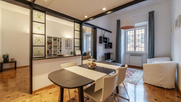 A dining area with a table, chairs, and a decorative bowl. There is a window with curtains and a radiator.