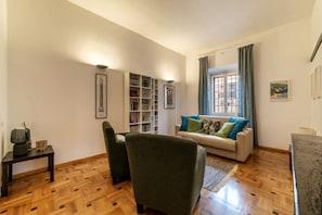 A living room with a sofa, armchairs, a coffee table, a bookshelf, and a window with curtains.