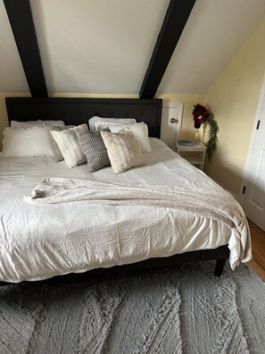 Un lit bien fait, avec une couette blanche et plusieurs oreillers, un tapis gris et une tête de lit sombre, dans une pièce au design simple et épuré.