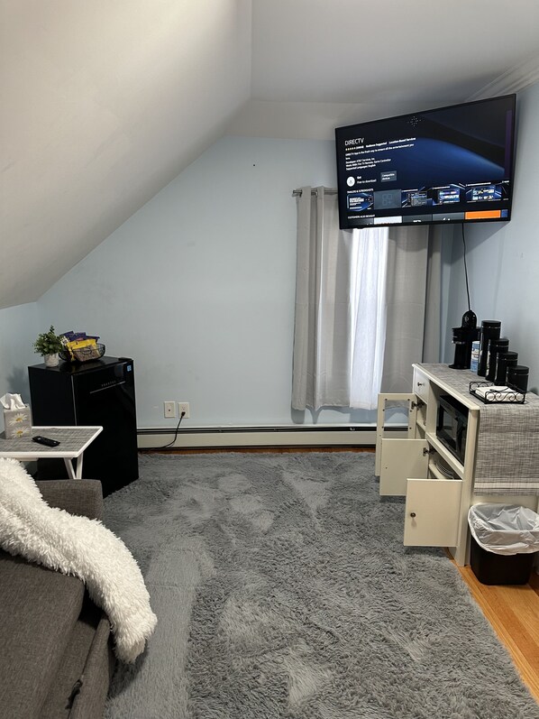 A small, sloped ceiling room with a flat-screen TV mounted on the wall, a small table with a remote control, a white fluffy throw blanket, a grey shaggy rug, and a white cabinet with a microwave. - Mini cozy quiet apartment (Manchester)