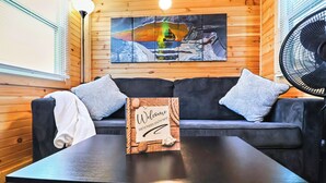 Um quarto aconchegante com parede de madeira, sofá preto com almofadas estampadas, um letreiro de boas-vindas e uma pintura de um barco na água.