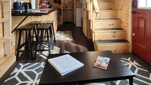 Uma cabana de madeira compacta com cozinha, mesa de jantar e cadeiras, e escada que leva a um andar superior.