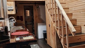 Interior de uma cabana de madeira com cozinha, uma mesa de jantar e escada que leva a um andar superior.