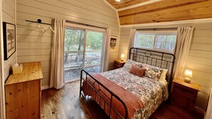 A bedroom with a wooden ceiling, a bed with floral bedding, a nightstand with a lamp, and a view through a large window.