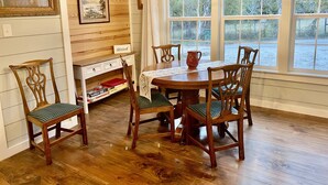 A dining area with a wooden table, six chairs, a chandelier, and large windows with a view of trees.