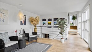 Un salon moderne avec un canapé, des fauteuils, une table basse et une grande plante en pot. Des œuvres d’art encadrées ornent le mur, et un escalier mène à l’étage supérieur.