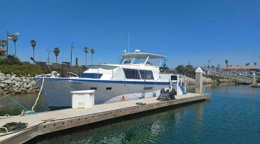 Un barco amarrado en un puerto deportivo, con cielo despejado y palmeras al fondo.