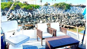 Una terraza con sillas blancas acolchadas y una mesa de madera, con vistas a una costa rocosa con palmeras.