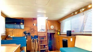Interior compacto de autocaravana con piso de madera, sofá con cojines azules, una mesita, un ventilador y una ventana con persianas.