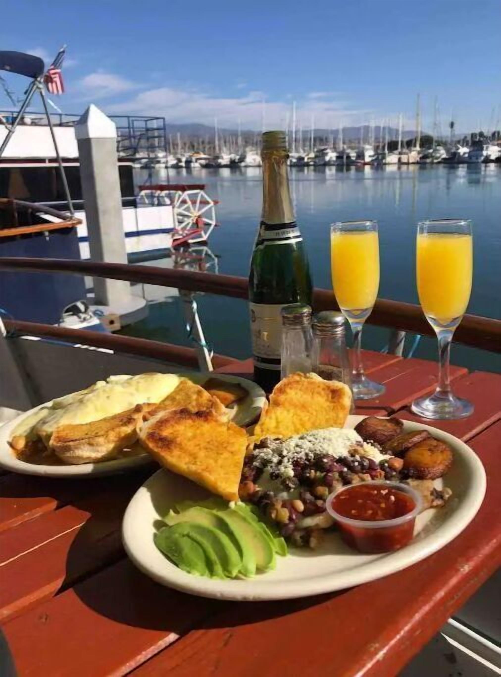 Een ontbijtbakje met toast, avocado, bonen, een klein schaaltje saus, een fles champagne en twee glaasjes jus d'orange op het dek van een boot, met uitzicht op een jachthaven.